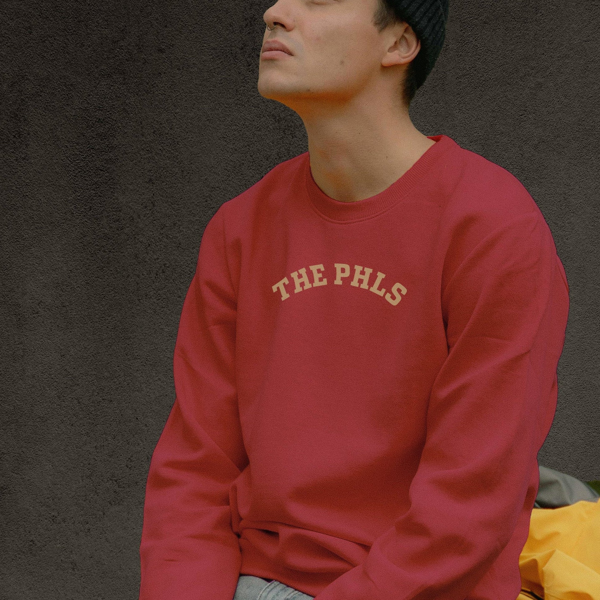 Person wearing a red sweatshirt with 'THE PHLS' text, sitting on a wooden crate against a dark background.