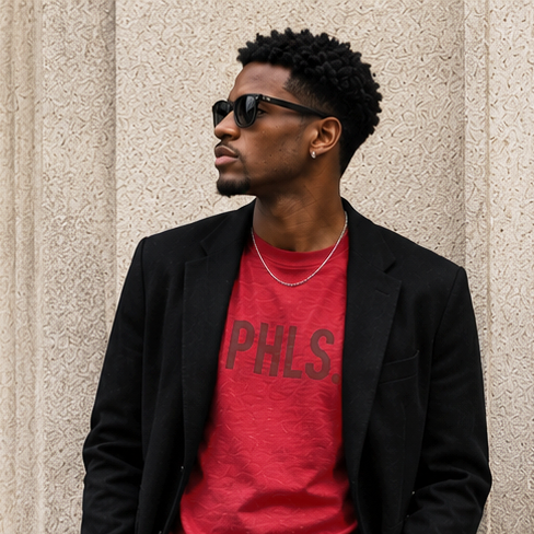 Man wearing a red 'PHLS' shirt and black jacket against a textured wall.