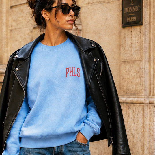 Woman in blue sweatshirt and jeans walking on a street, wearing sunglasses and carrying a black handbag.