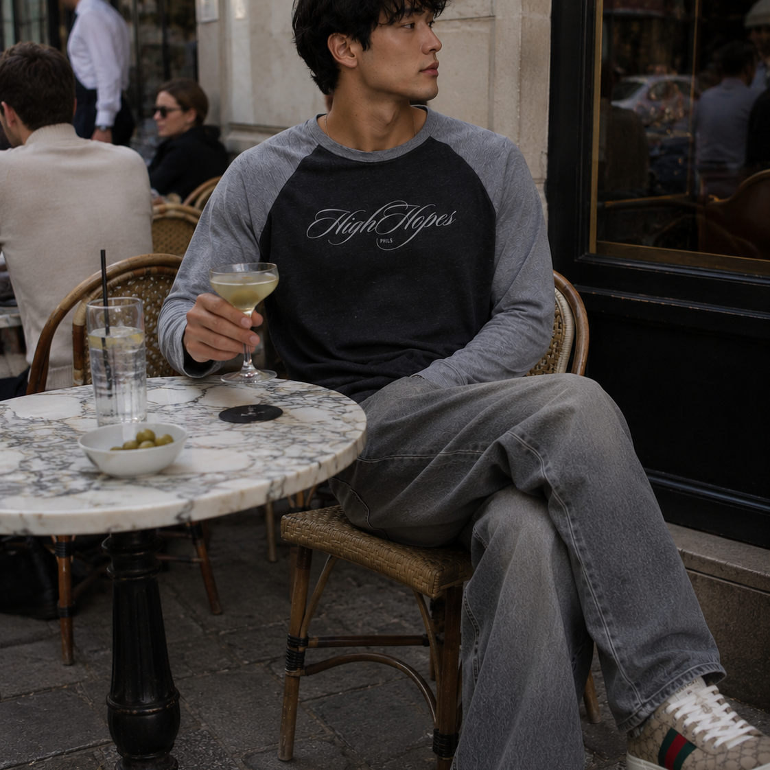 Man sitting at an outdoor cafe holding a drink, wearing a black and gray sweatshirt with text.