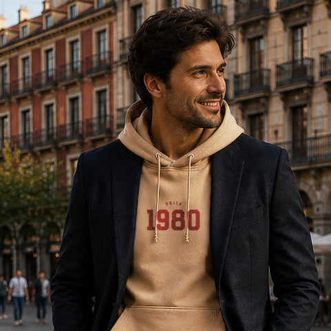 Man wearing a beige hoodie with '1980' print, black jacket, and white sneakers in an urban setting.