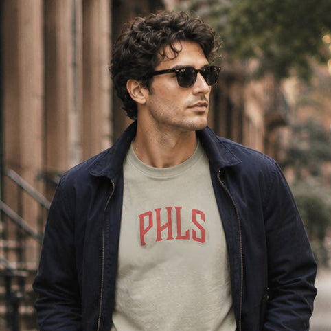 Man walking on a sidewalk wearing a gray 'PHILS' t-shirt, navy jacket, green pants, and white sneakers.