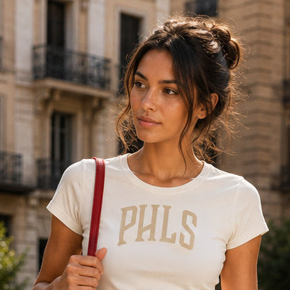 Woman wearing a white t-shirt with 'PHLS' text, holding a red bag in an urban setting.