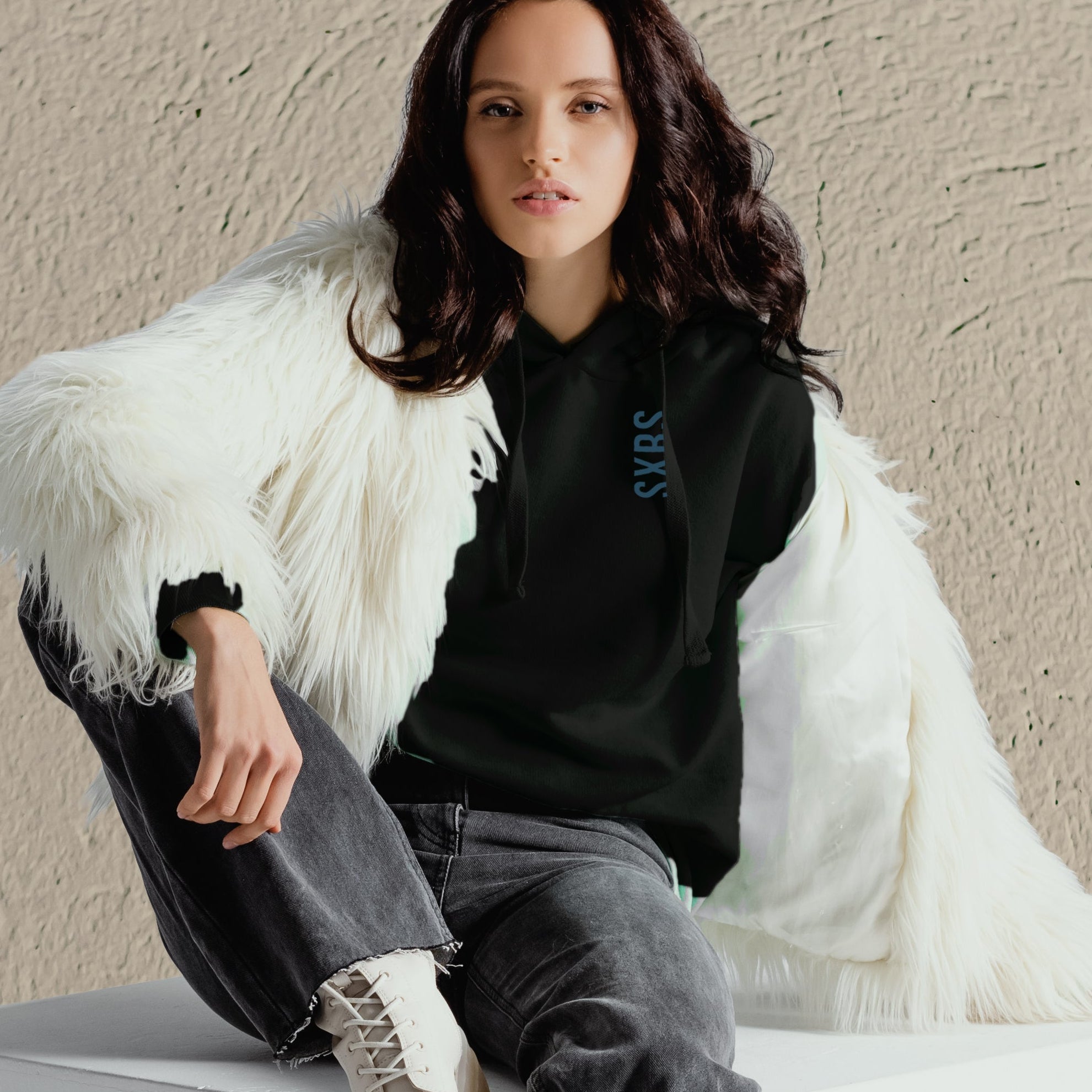 Woman wearing a black hoodie with a logo, gray pants, and white sneakers, sitting against a beige wall.