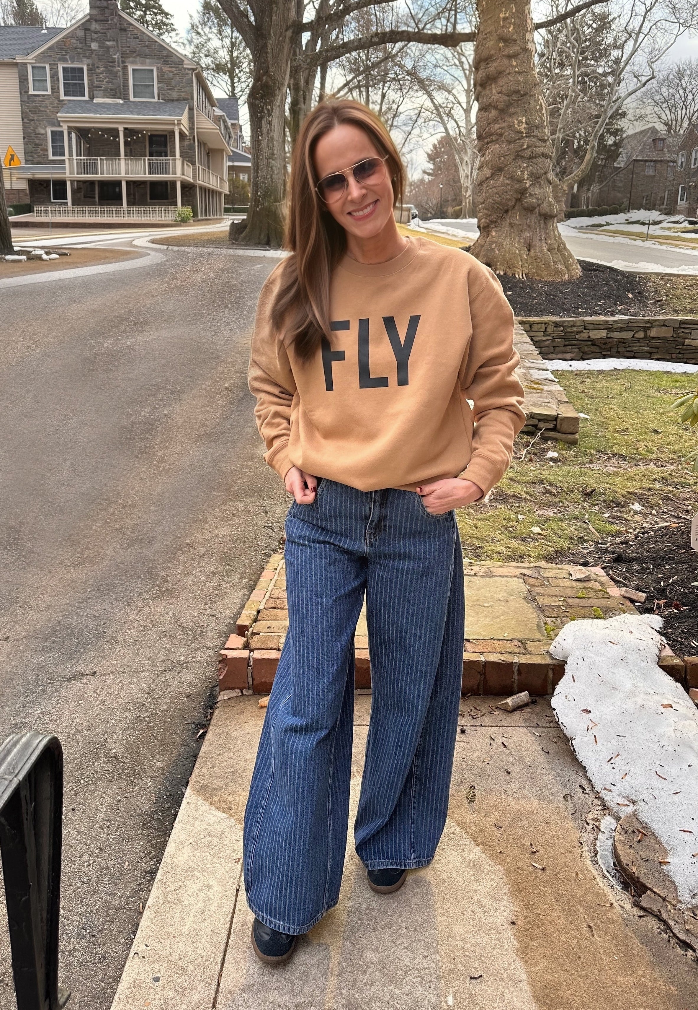 Woman wearing a beige sweatshirt with 'FLY' text and blue jeans standing on a sidewalk.