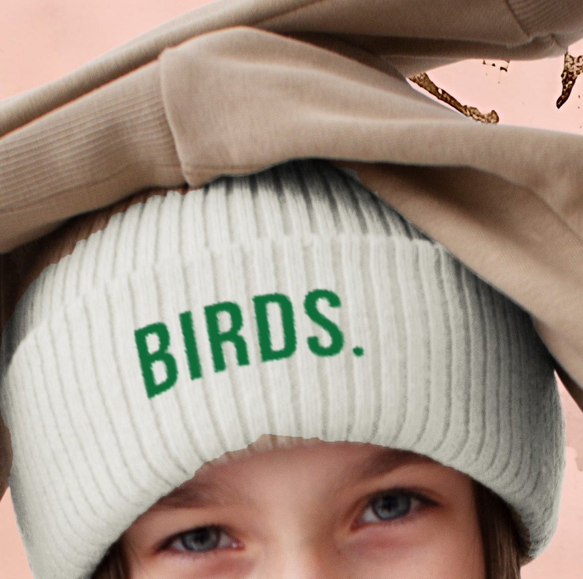 Child wearing a white beanie with 'BIRDS.' text, holding a beige coat over their head against a pink background