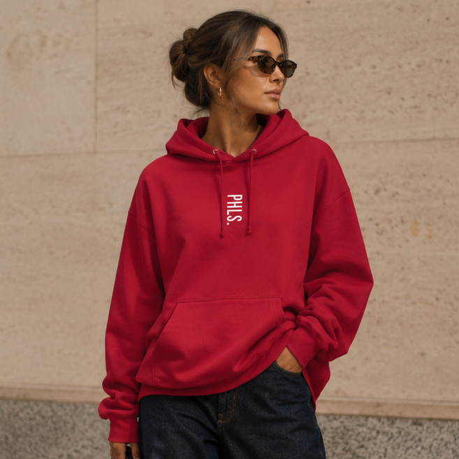 Woman wearing a red hoodie and black pants, holding a black handbag, standing against a beige building.