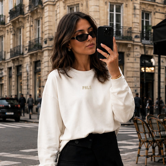 Woman taking a selfie on a city street, wearing a white sweatshirt and black skirt.
