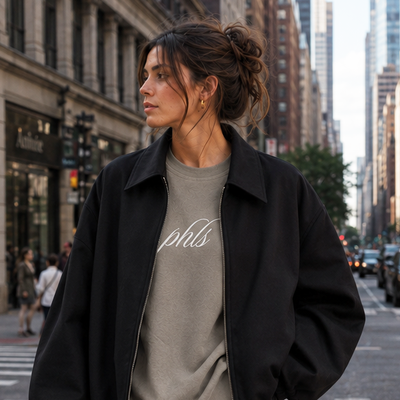 Woman standing on a city street wearing a black jacket, gray sweatshirt, and denim shorts.