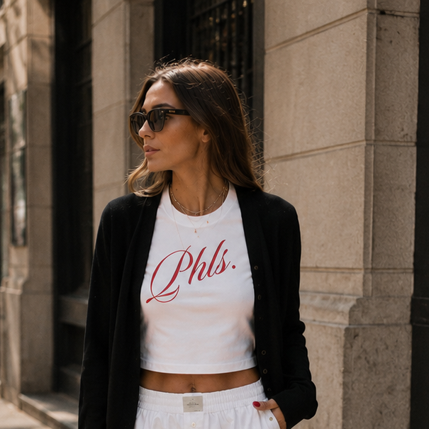 Woman walking on a city street wearing a white crop top with text, black blazer, white shorts, and black shoes.