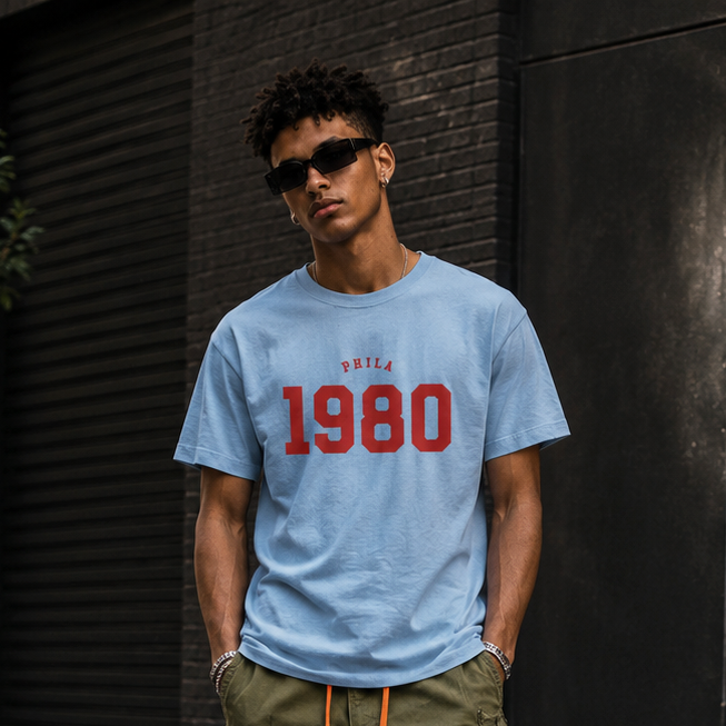 Person wearing a light blue t-shirt with '1980' printed on it, green cargo pants, and black sneakers, standing against a dark wall.