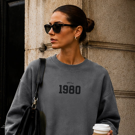 Woman walking outdoors holding a coffee cup, wearing a gray sweatshirt with '1980' text.
