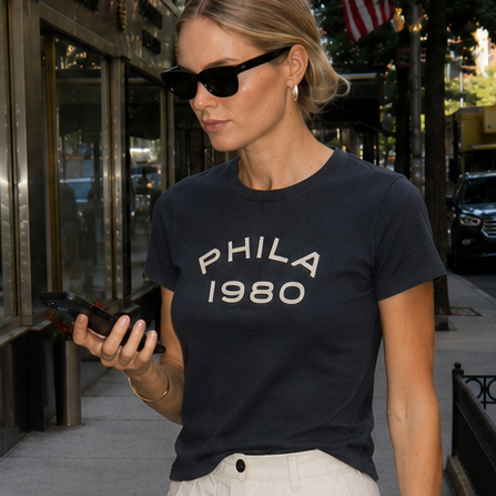 Woman in a black t-shirt with 'PHILA 1980' text, white shorts, and sunglasses standing on a city street.