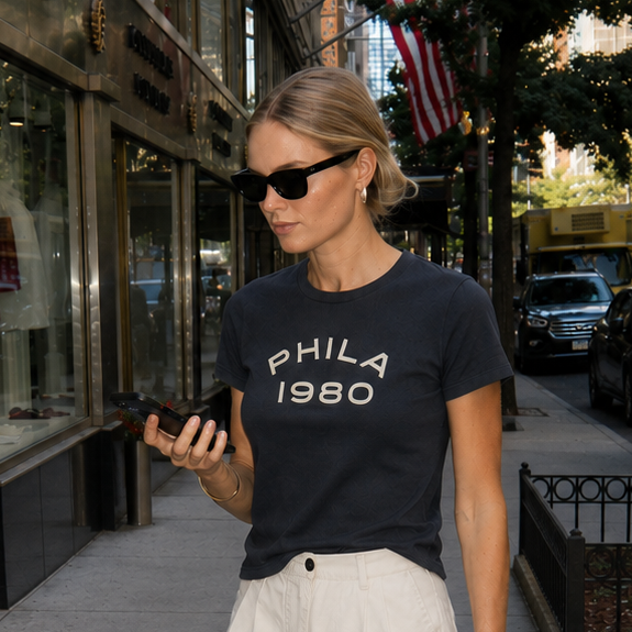 Woman in a black t-shirt with 'PHILA 1980' text, white shorts, and sunglasses standing on a city street.