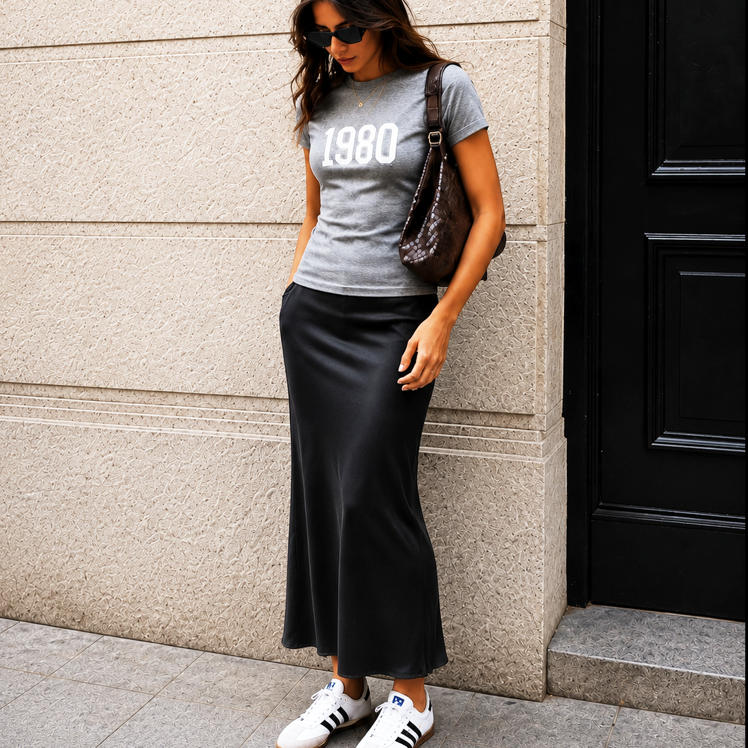 Woman wearing a gray t-shirt with '1980' text, black skirt, and white sneakers against a building.