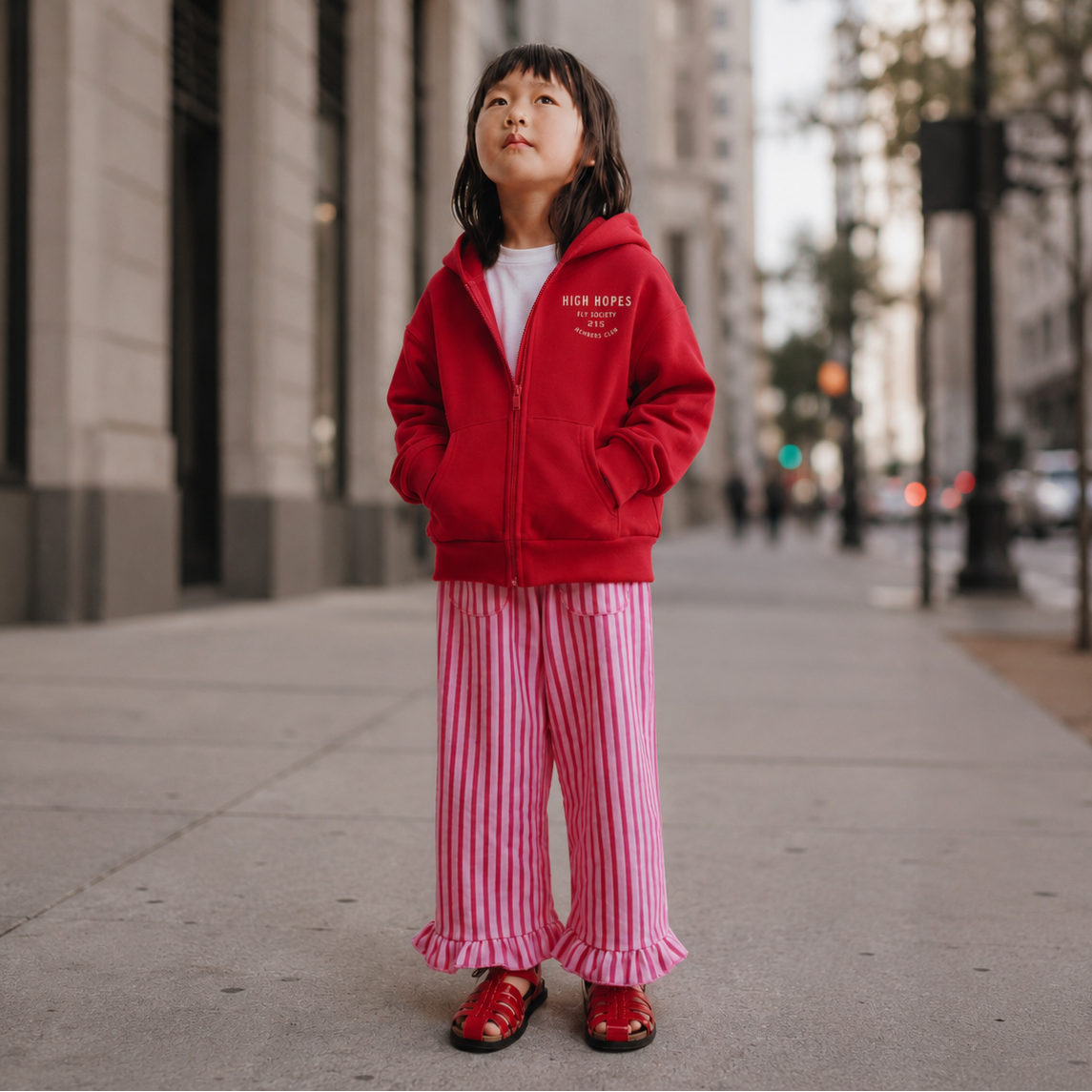 Child wearing a red jacket and pink striped pants standing on a city street.
