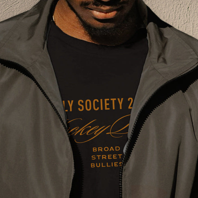 Person wearing a dark jacket and black t-shirt with text, holding a phone against a neutral background